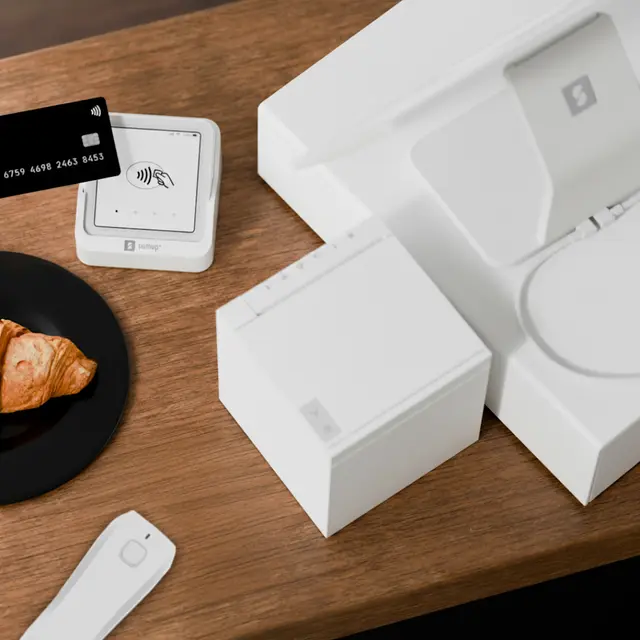 White SumUp POS hardware on a wooden countertop with card reader, receipt printer, and display stand beside a black payment card and croissant