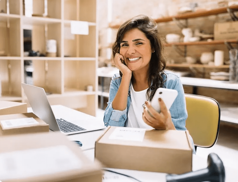 Smiling small business owner holding a smartphone at a desk with parcels and a laptop in the background