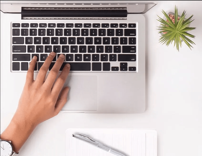 Top-down view of a hand typing on a laptop keyboard beside a notebook, pen, and small plant