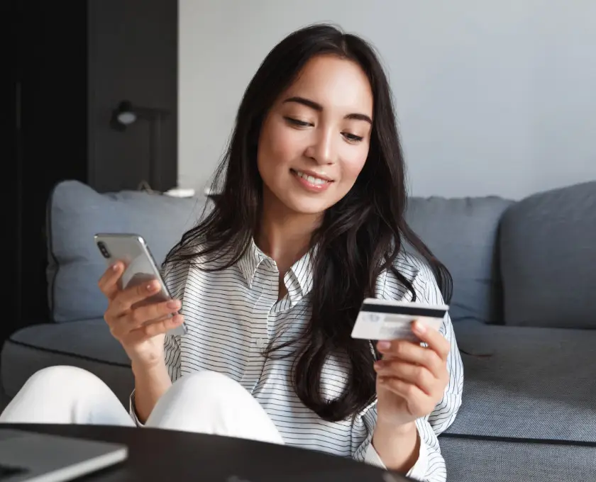 Woman shopping online at home while holding a smartphone and payment card on a sofa